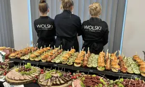 Trzy kobiety w czarnych fartuchach z logo "WOLSKI CATERING" stoją za stołem pełnym przekąsek. To idealna impreza z pysznym jedzeniem.