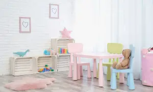 Kolorowy stolik z krzesełkami dla dzieci, idealny do zabawy i nauki. Na ścianie wiszą ramki z serduszkami, a obok stoją drewniane skrzynki z zabawkami.
