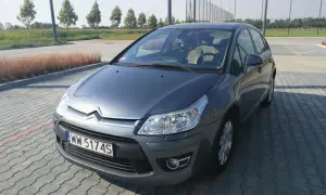 Szary Citroen C4 z 2008 roku na parkingu z kostki brukowej, w tle drzewa i boisko.