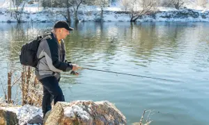 Mann angelt bei Minusgraden am Flussufer, das mit Schnee bedeckt ist. Er trägt eine Jacke und einen Rucksack.