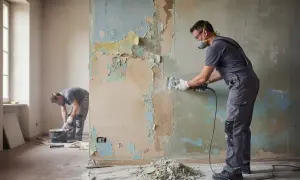 Uomo con maschera e occhiali esegue la rasatura di un muro scrostato con una levigatrice.