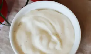 Biały sos śmietanowy w miseczce, z widocznymi drobinkami pieprzu, na tle pomidorów.
