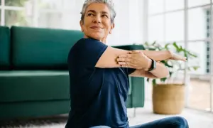 Une femme souriante s'étire, prouvant qu'il est possible de redresser son dos à 60 ans. Elle est assise sur un tapis, près d'un canapé vert.