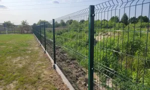 Nowe, zielone ogrodzenie działki rekreacyjnej z betonowym podmurowaniem. W tle trawa i zarośla.