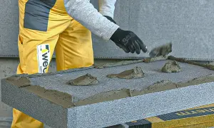 Nakładanie kleju do styropianu grafitowego na płytę izolacyjną. Ręce w czarnych rękawiczkach rozprowadzają masę szpachlą.