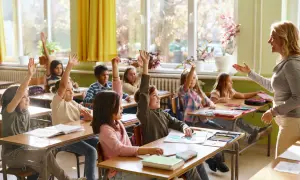 Grundschüler mit erhobenen Händen, bereit zu antworten. Die Sitzordnung zur Wand hin ist gut erkennbar, die Lehrerin lächelt.