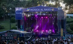 Escenario iluminado con luces púrpuras para un concierto de **música al raso**. Una multitud disfruta del espectáculo al aire libre.