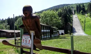 Drewniana figura skoczka narciarskiego przed skocznią w Harrachovie. W tle zielone wzgórza i lasy.