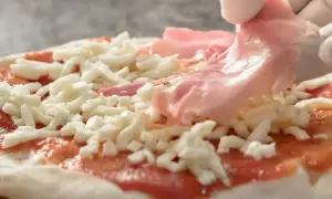 Rękawiczka w białej rękawiczce dodaje cienkie plastry różowej szynki na pizzę, która jest już pokryta sosem pomidorowym i startym serem.