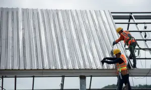 Dwóch budowlańców w kaskach i kamizelkach montuje dach z blachy trapezowej, pokazując, jak wkręcać wkręty do blachy trapezowej.