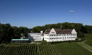 Schloss Hersberg, umgeben von Weinbergen, mit einem Zeppelin am blauen Himmel.