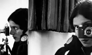 Tina Modotti w kadrze, z aparatem w ręku, patrzy w obiektyw.