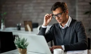 Mężczyzna w okularach, skupiony na laptopie, pracuje przy biurku. W tle widać elementy biurowe, tworząc atmosferę miejsca pracy. Ten sygnalista w pracy analizuje dane.