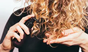 Delikatna pielęgnacja włosów wysokoporowatych. Ręka z buteleczką kosmetyku aplikuje go na blond loki.