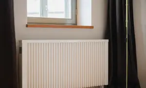 Biały ekran zagrzejnikowy stoi przy oknie, obok ciemnej zasłony i stojącej lampy. Idealny do wnętrz, gdzie liczy się estetyka i funkcjonalność.
