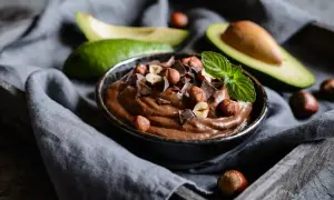 Kremowa nutella z awokado, udekorowana orzechami laskowymi, kawałkami czekolady i miętą. Obok leżą połówki awokado.