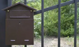 Brązowa skrzynka na listy w kształcie domku, zamontowana na czarnym słupku ogrodzenia. Zamykana na kluczyk, z miejscem na nazwisko.