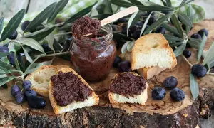 Ecco come si fa il patè di olive: bruschette con paté scuro, olive nere e rametti d'ulivo su un tagliere di legno.