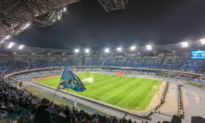 Widok na pełen kibiców stadion w Neapolu nocą. Trybuny wypełnione niebieskimi barwami, na murawie zielona trawa.