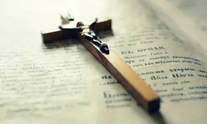 Drewniany krzyż z Jezusem na otwartej księdze. Tekst na stronach przypomina modlitwy lub fragmenty Pisma Świętego, być może związane z płaszczem św. Józefa.