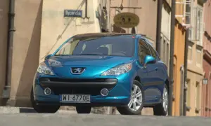 Błękitny Peugeot 207 z wyraźnymi żarówkami reflektorów stoi na brukowanej ulicy.
