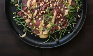 Rinder Carpaccio mit Pinienkernen, Kapern und Rucola auf schwarzer Platte, beträufelt mit einer cremigen Sauce.