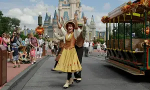 Uczestnicy parady w Disneylandzie pozdrawiają tłum. W tle zamek, a obok zabytkowy tramwaj.