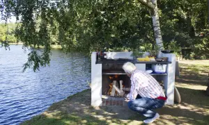 Man tänder en **mura grill** vid en sjö. Elden sprakar och förberedelser för grillning pågår.