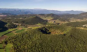 Paisaje de la ruta volcanes Garrotxa, con conos volcánicos cubiertos de bosque y campos verdes, bajo un cielo azul.