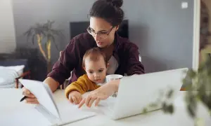 Kobieta z dzieckiem na kolanach pracuje przy laptopie, przeglądając notatki. Zastanawia się, ile zarabia opiekunka do dziecka, gdy maluch próbuje jej pomóc.
