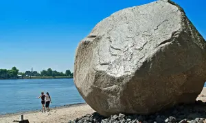 Der alte Schwede, ein riesiger Findling, thront am Ufer. Zwei Frauen spazieren am Wasser entlang, im Hintergrund Bäume und ein blauer Himmel.