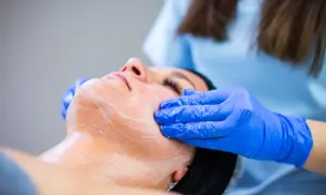 Zabieg oxybrazji: kosmetyczka w niebieskich rękawiczkach delikatnie masuje twarz klientki pokrytą pianką.
