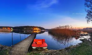 Czerwona łódka czeka przy pomoście na Jeziorze Skępe. W tle nocne światła lasu i niebo pełne gwiazd.