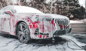 Czerwony samochód pokryty pianą podczas mycia. Widać felgi, reflektory i grill. W tle drzewa i schody.