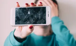 Złamany telefon z pękniętym ekranem, który osoba trzyma przed twarzą, zakrywając ją ręką.