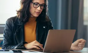 Kobieta w okularach i skórzanej kurtce pracuje na laptopie, szukając informacji, jak sprawdzić lata pracy w ZUS PUE.