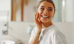 Une femme souriante applique une crème sur sa joue. Elle se prépare pour la journée, peut-être un primer avant ou après crème solaire.
