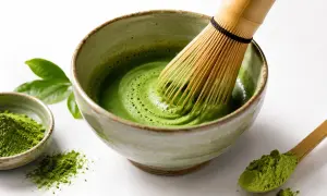 Tradycyjny matcha przepis: bambusowa miotełka ubija zielony proszek w misce, tworząc piankę. Obok miseczka z proszkiem i drewniana łyżeczka.