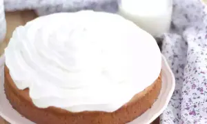 En saftig tres leches-kaka med fluffigt vitt täcke, bredvid ett glas mjölk och en blommig duk.