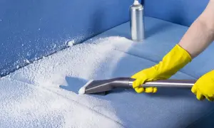Person in yellow gloves using a vacuum attachment to clean a blue fabric sofa with white foam.
