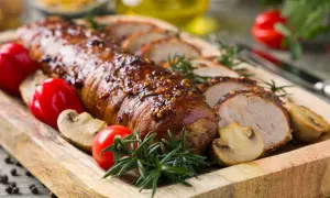 Lomo de cerdo glaseado con mostaza, servido con champiñones, tomates y romero en una tabla de madera.
