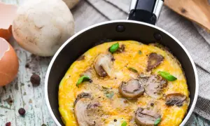 Pyszny omlet z pieczarkami, świeżo usmażony na patelni, z dodatkiem bazylii. Obok skorupki jajka i grzyby.