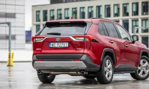 Czerwony Toyota RAV4 2020 Hybrid na mokrym parkingu, z nowoczesnym budynkiem w tle.