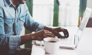 Mężczyzna w dżinsowej koszuli pisze na laptopie, obok stoi filiżanka kawy.