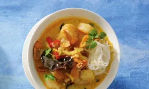 Aromatyczna zupa krewetkowa po tajsku z krewetkami, makaronem ryżowym, papryczką chili i kolendrą na niebieskim tle.