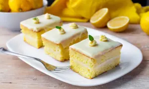 Tre fette di torta al limone e cioccolato bianco, guarnite con glassa e foglioline verdi, su un piatto bianco con limoni sullo sfondo.