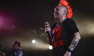 Wokalista punk rockowy z irokezem śmieje się do mikrofonu. Na koszulce ma czerwony nadruk.