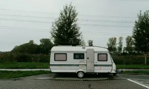 Ein weißer b96 wohnwagen steht auf einem gepflasterten Parkplatz, umgeben von grüner Wiese und Bäumen unter einem grauen Himmel.