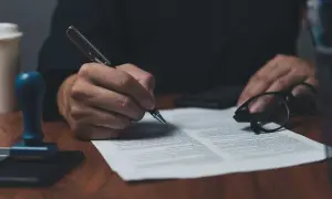 Dłoń podpisuje dokument piórem, obok okulary i pieczęć. To ważny list żelazny, potwierdzający umowę.