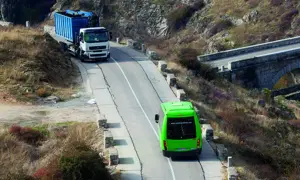 Camión blanco con contenedor azul subiendo una pendiente, seguido por un autobús verde. Se debate quién tiene prioridad en una pendiente.